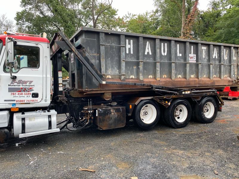 Roll-off truck carrying a steel Haul It All dumpster, parked on a gravel lot