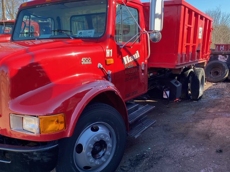 Red roll-off truck with a red dumpster loaded on the back, parked on a dirt lot